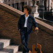 A person stands on outdoor stone steps beside a brick wall, wearing a dark navy Shacket and matching trousers over a white shirt, with a brown acoustic guitar resting beside them. Historic buildings and statues are visible in the background.