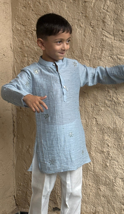 A young boy modeling a light blue long-sleeve Mal Cotton kurta with white trousers and gray slip-on shoes, standing against a textured beige plaster wall.