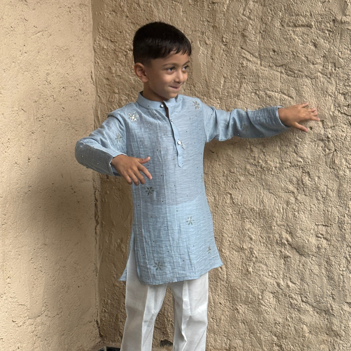 A young boy modeling a light blue long-sleeve Mal Cotton kurta with white trousers and gray slip-on shoes, standing against a textured beige plaster wall.