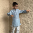 A young boy modeling a light blue long-sleeve Mal Cotton kurta with white trousers and gray slip-on shoes, standing against a textured beige plaster wall.