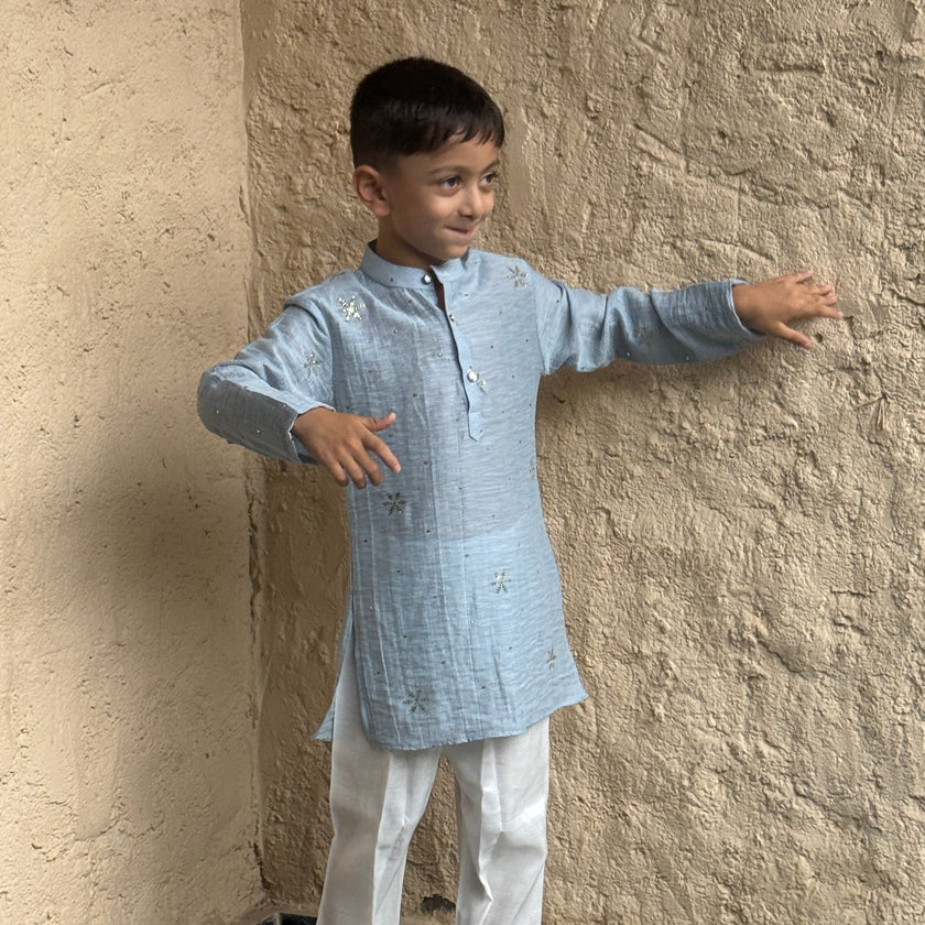 A young boy modeling a light blue long-sleeve Mal Cotton kurta with white trousers and gray slip-on shoes, standing against a textured beige plaster wall.