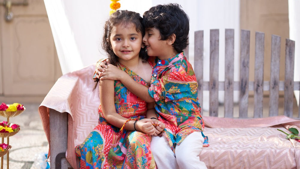 A young girl and boy sit closely together on a decorated pink bed in a traditional indoor setting. The girl wears a colorful patterned pink and blue dress, and the boy wears a matching colorful printed kurta with white pants.
