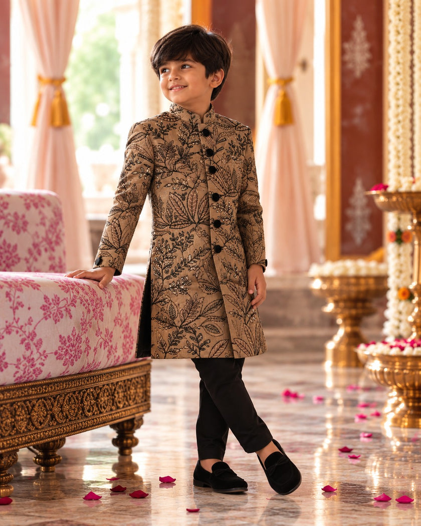 Boy leaning on a palace sofa in the long-line camel brown embroidered achkan and black pants