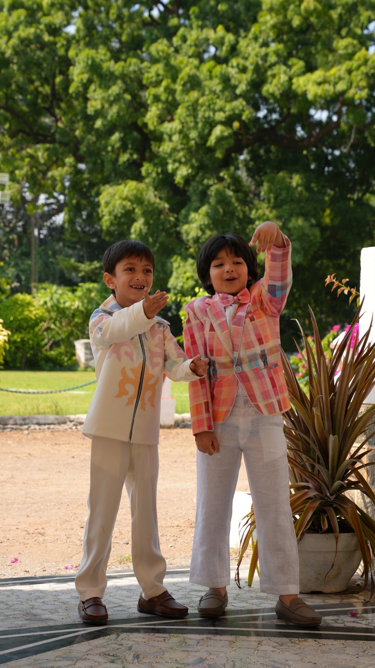 Two boys dancing outdoors — one in the ivory pastel appliqué zipper jacket and pant set, the other in a pink check blazer